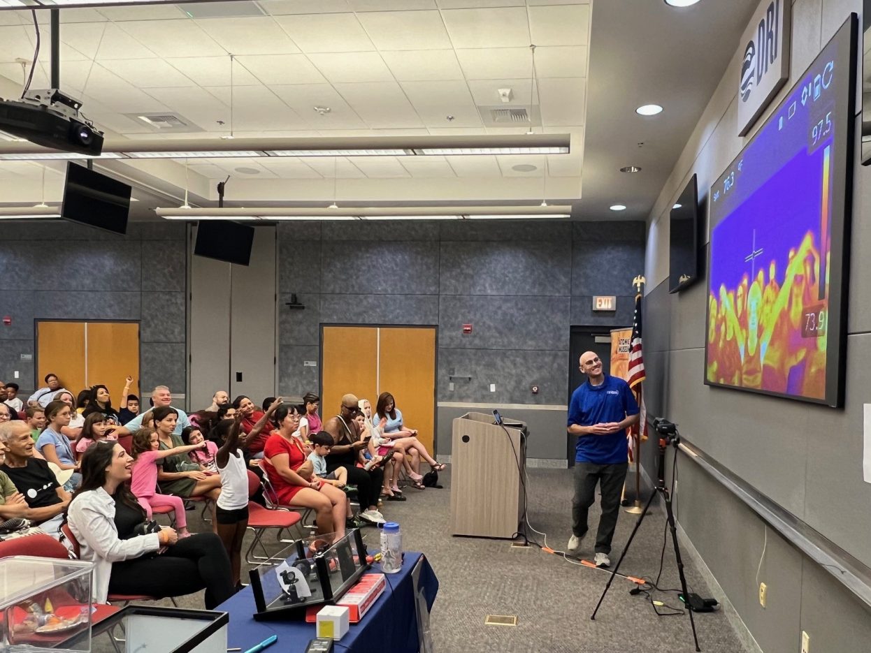 A NNSS scientist demonstrates an infrared camera as dozens of audience members point and smile.