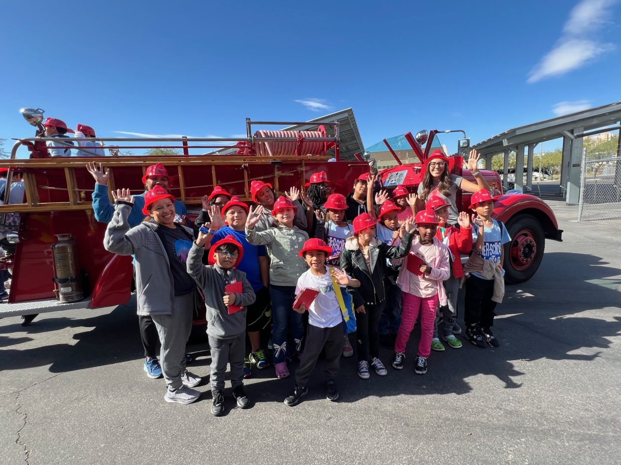 A group of students wear red firefighter hats while waving.