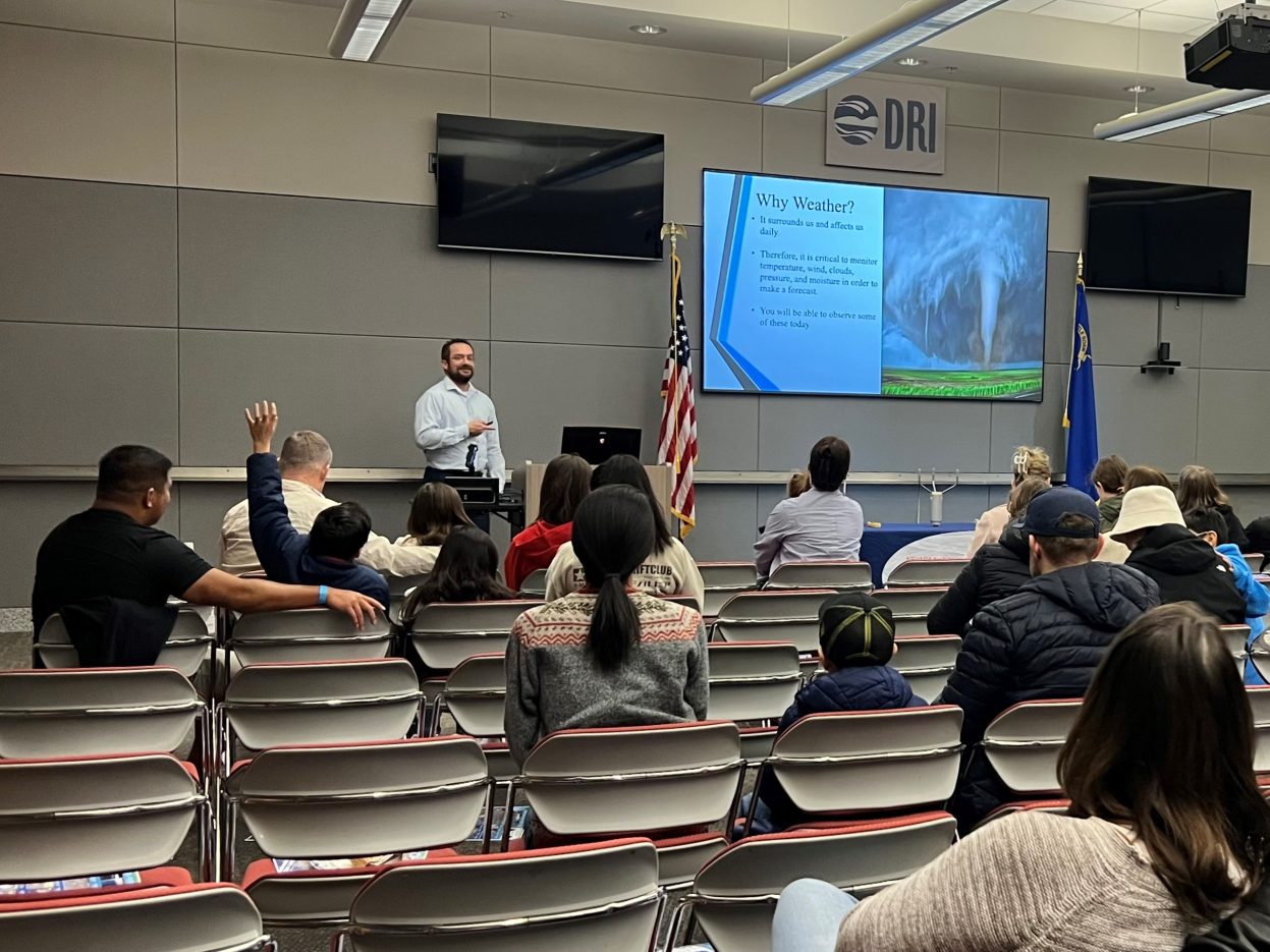 A meteorologist presents to a crowd, while one student raises his hand.