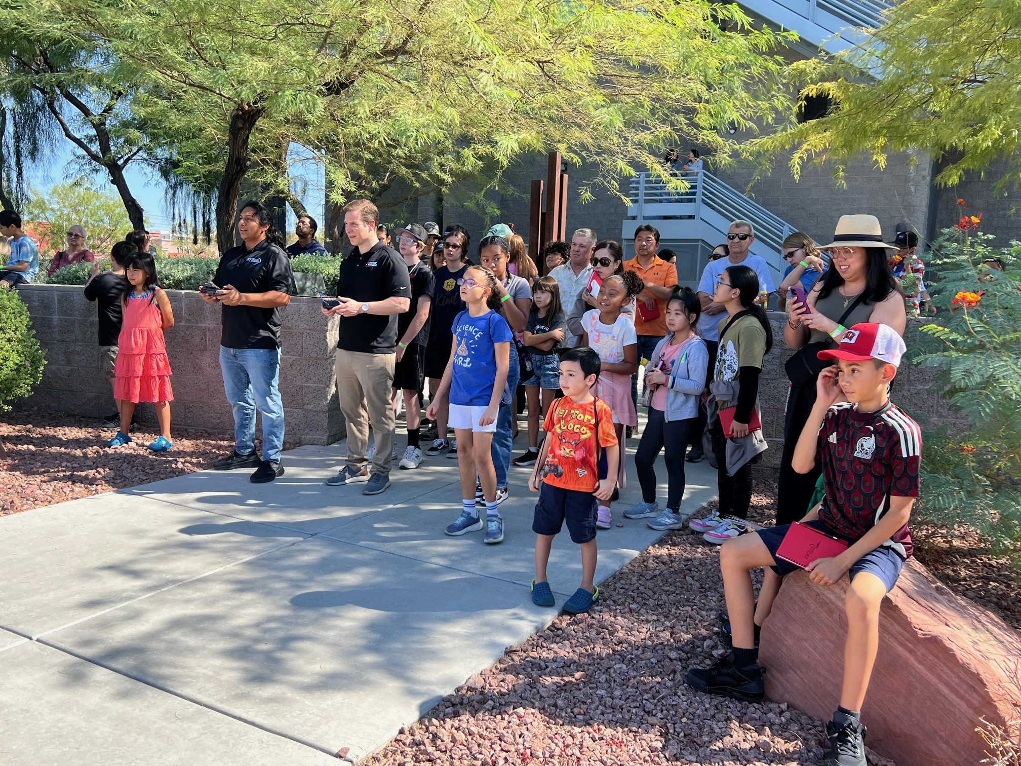 A group of students and parents watch a drone operate outdoors.