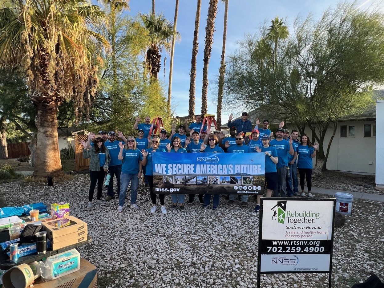 NNSS volunteers in blue-t-shirts wave from the front yard of a property. 