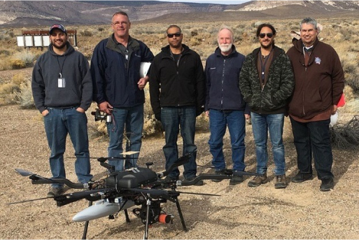 group of six men standing in desert with UAV on ground in front of them