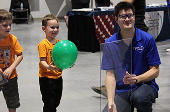two young children, one holding green balloon, with NNSS scientist holding up pane of glass