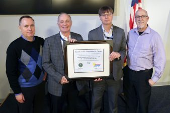 four men holding framed certificate with American flag in background on right side