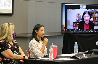 two women at table and one showing on TV screen in a conference room