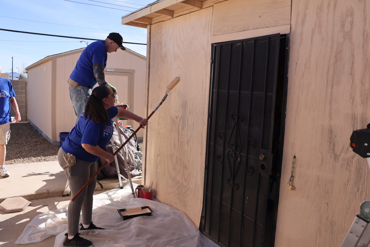 man in blue shirt on a ladder and women in blue shirt rolling paint on a shed