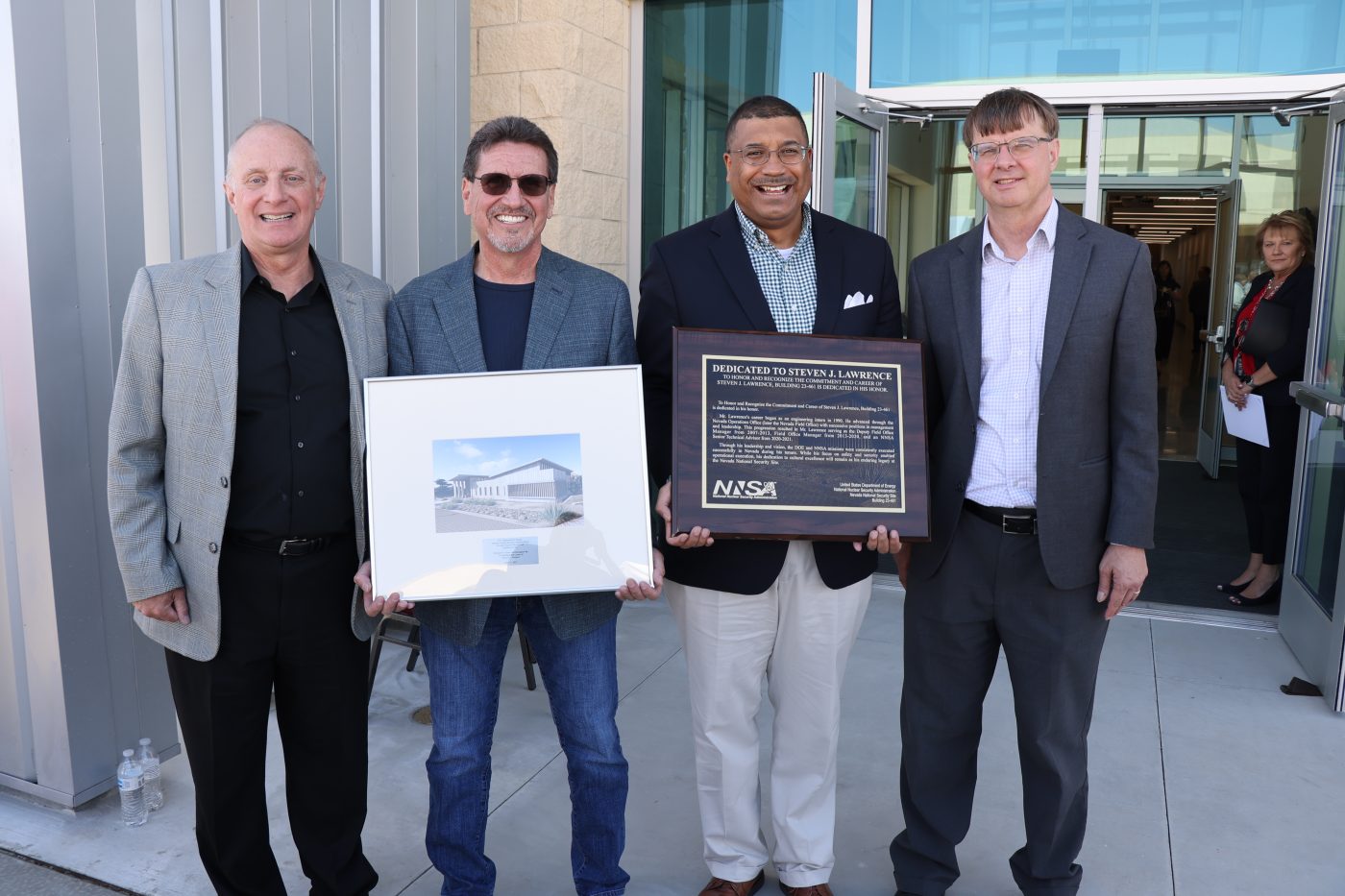 Four men in suit jackets standing in front of a new building with the two in the center holding a framed photo and a plaque