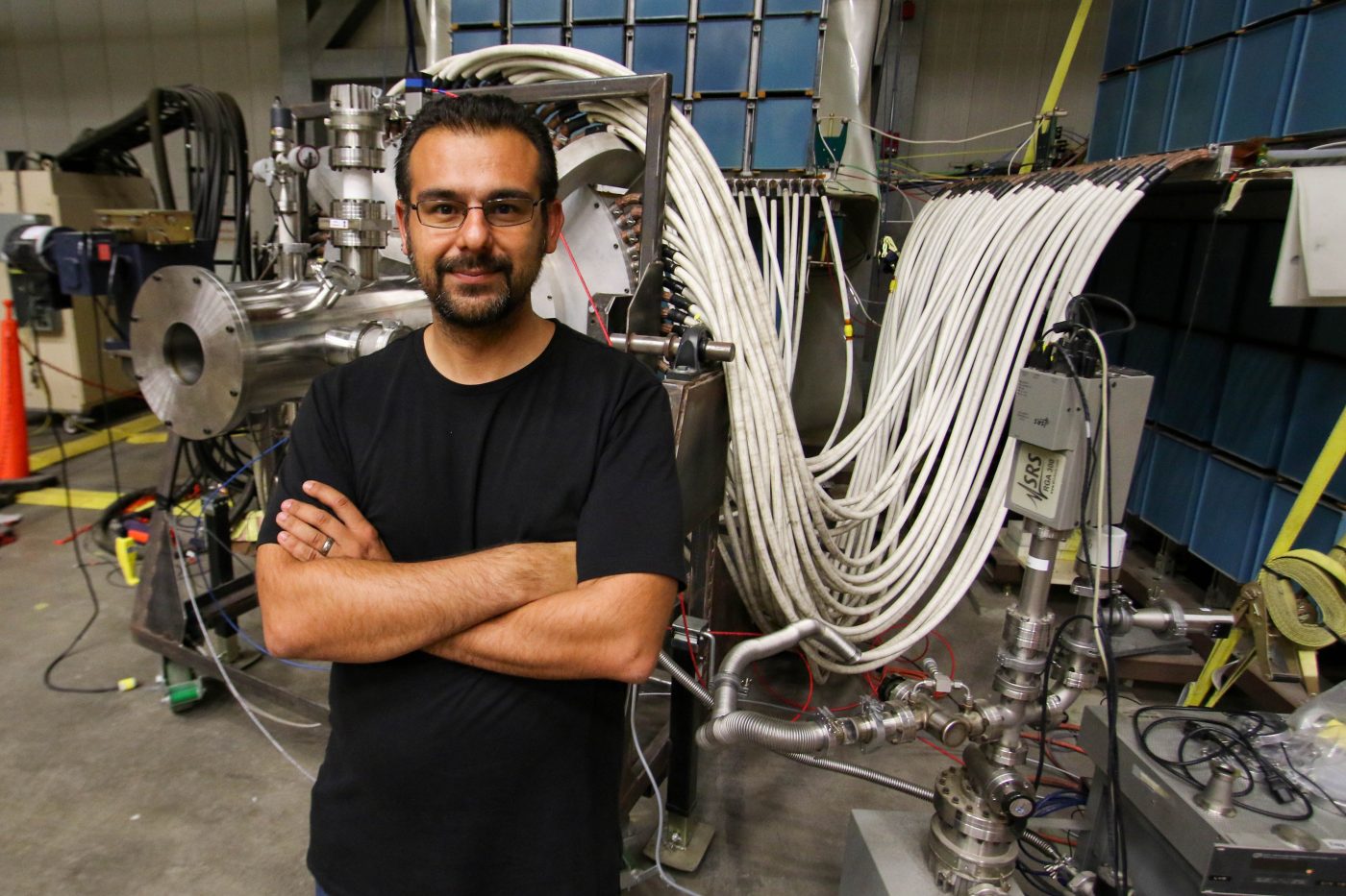 man in black t-shirt in forefront with glasses and dark hair and arms folded standing in front of dense plasma focus machine showing numerous white cables