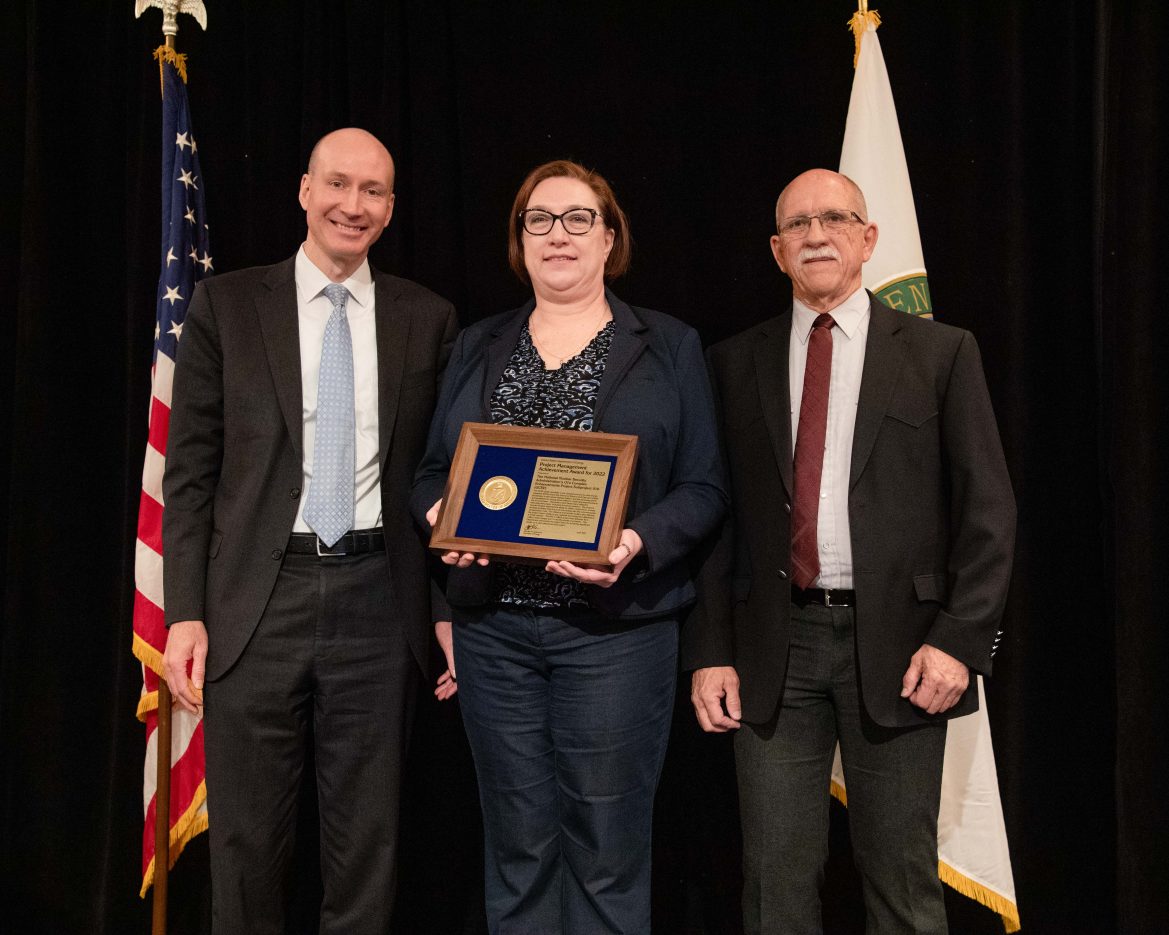 woman in middle holds framed award with men on either side of her in front of black background and American and DOE flags