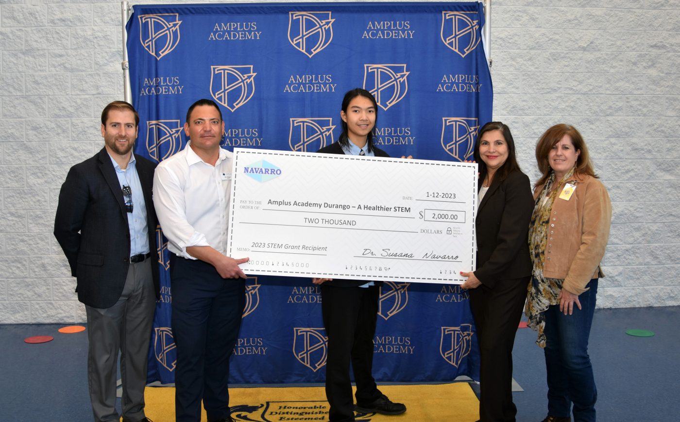 small group of children and adults holding big check in front of a blue backdrop with school logos on it