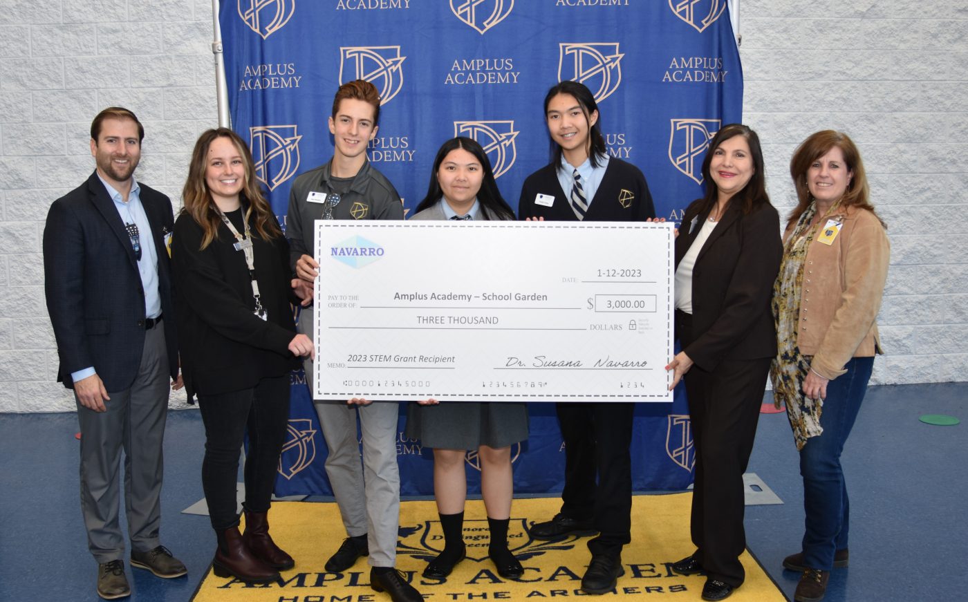 small group of children and adults holding big check in front of a blue backdrop with school logos on it