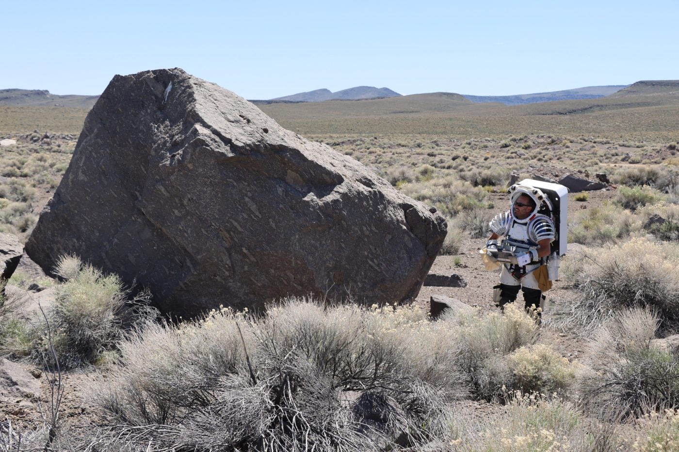 male astronaut candidate in white space suit standing in the desert next to a giant rock