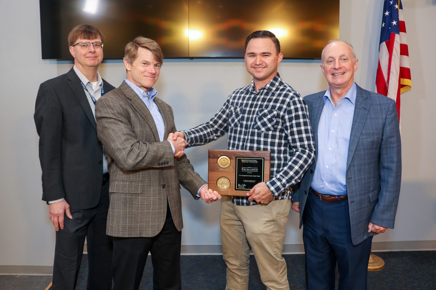 four men on stage with one in center accepting plaque and shaking hands