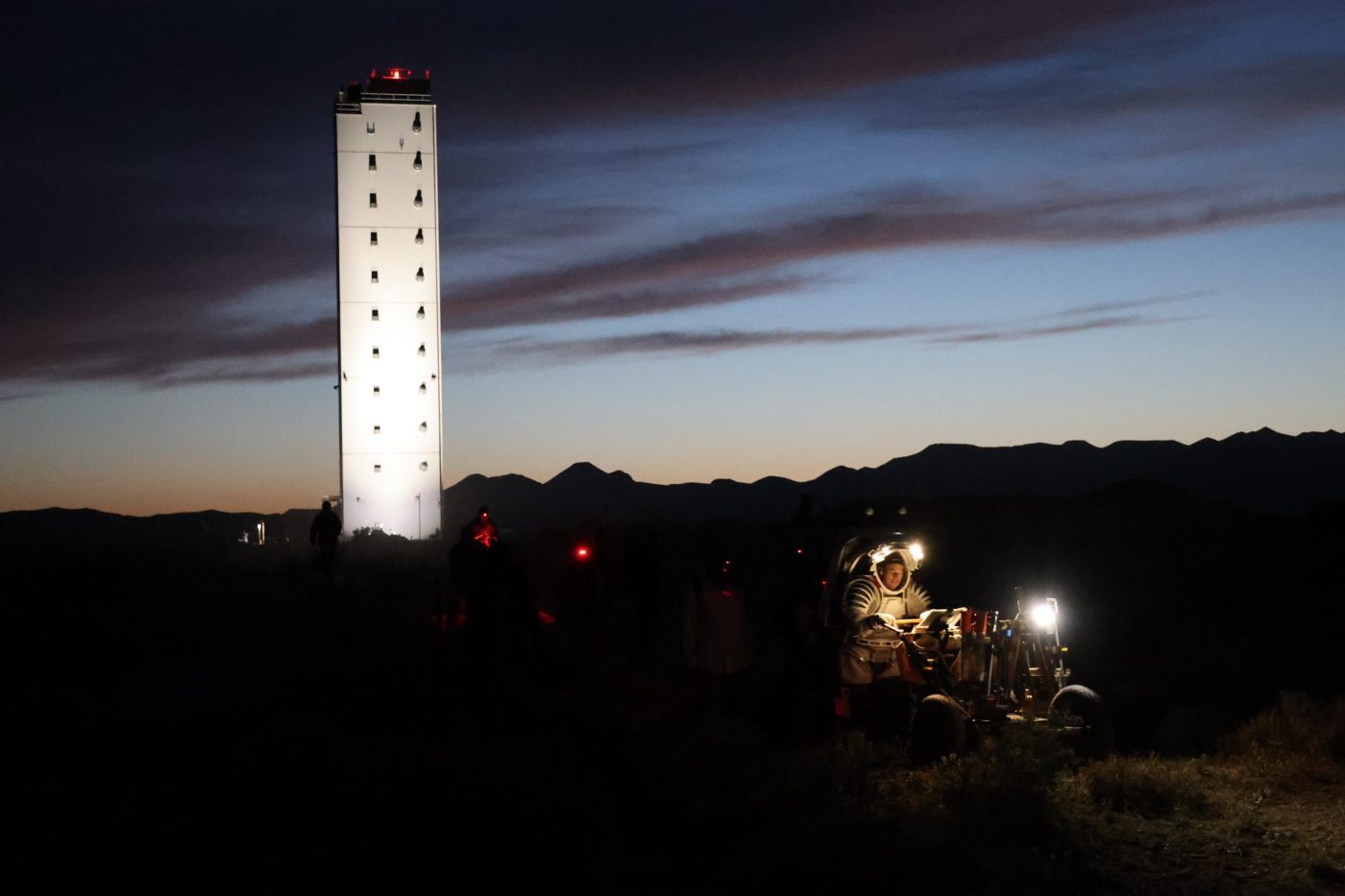 nighttime photo with tall white tower in background and NASA scientist in suit pushing a small vehicle both with headlamps on