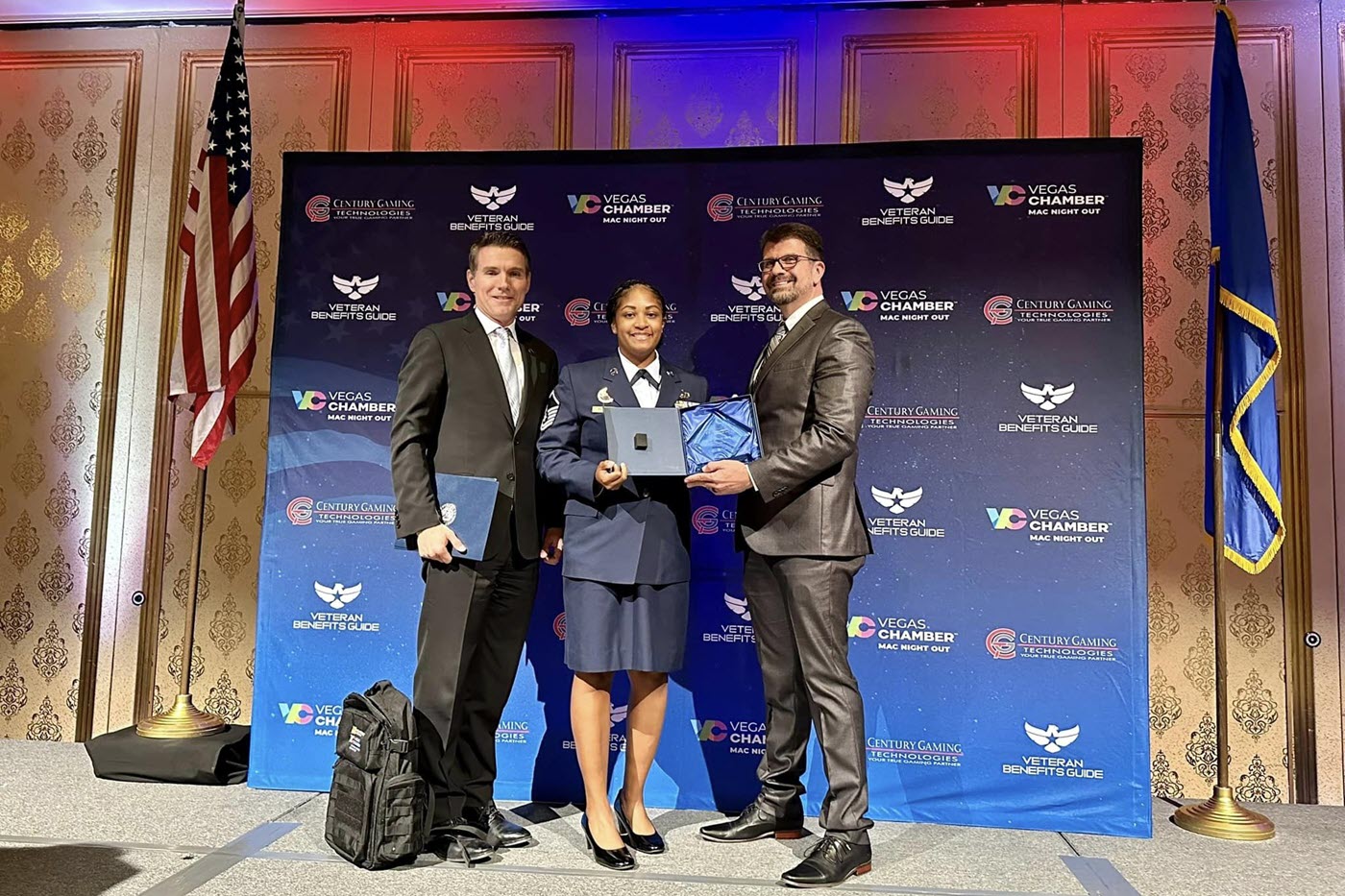 man on left in black suit, woman in center in Air Force dress blues, and man on right in dark gray suit holding glass award on stage in front of flags and backdrop during awards ceremony