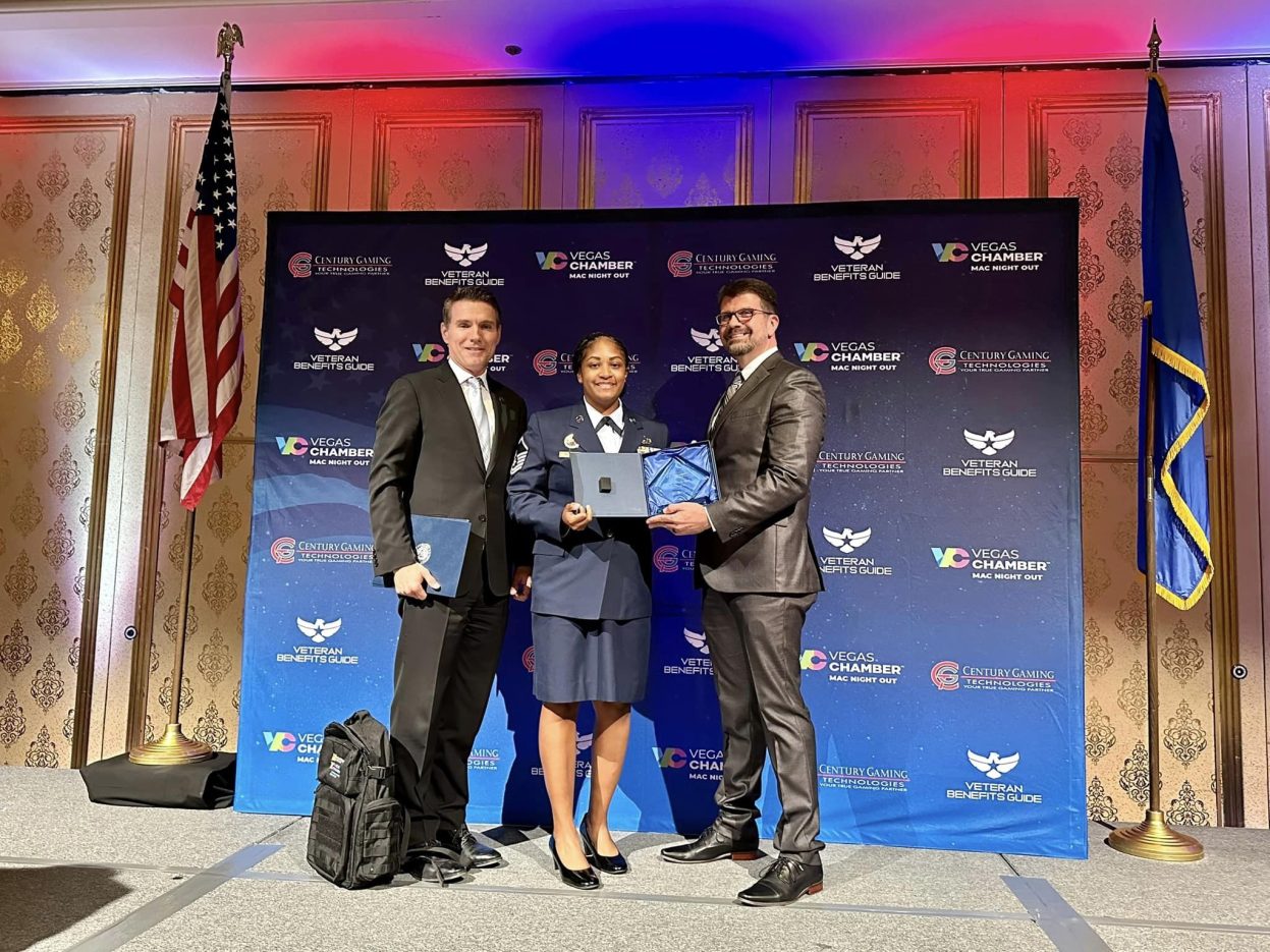 man on left in black suit, woman in center in Air Force dress blues, and man on right in dark gray suit holding glass award on stage in front of flags and backdrop during awards ceremony