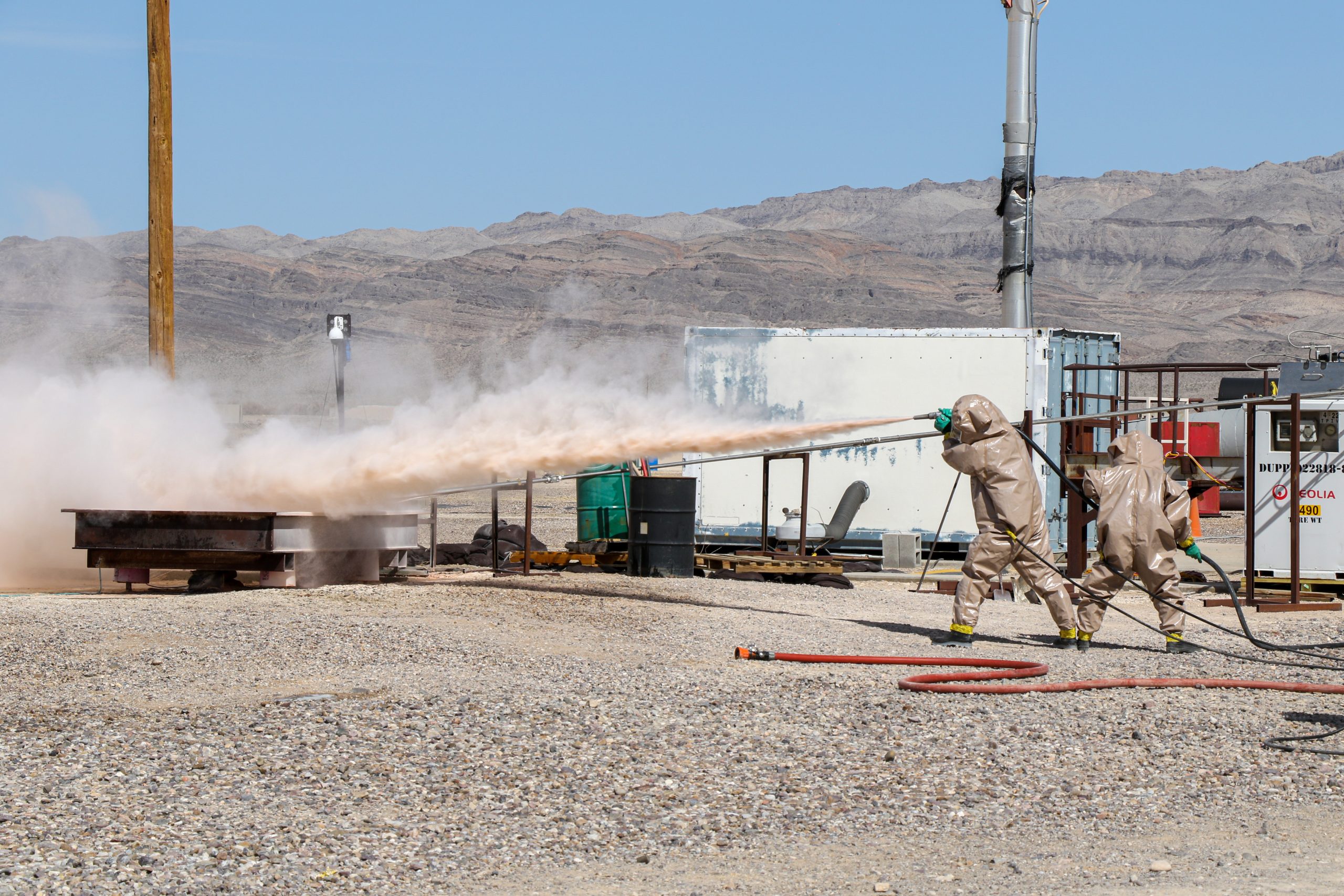 two people in brown protective coveralls spraying foam from a hose at a metal platform