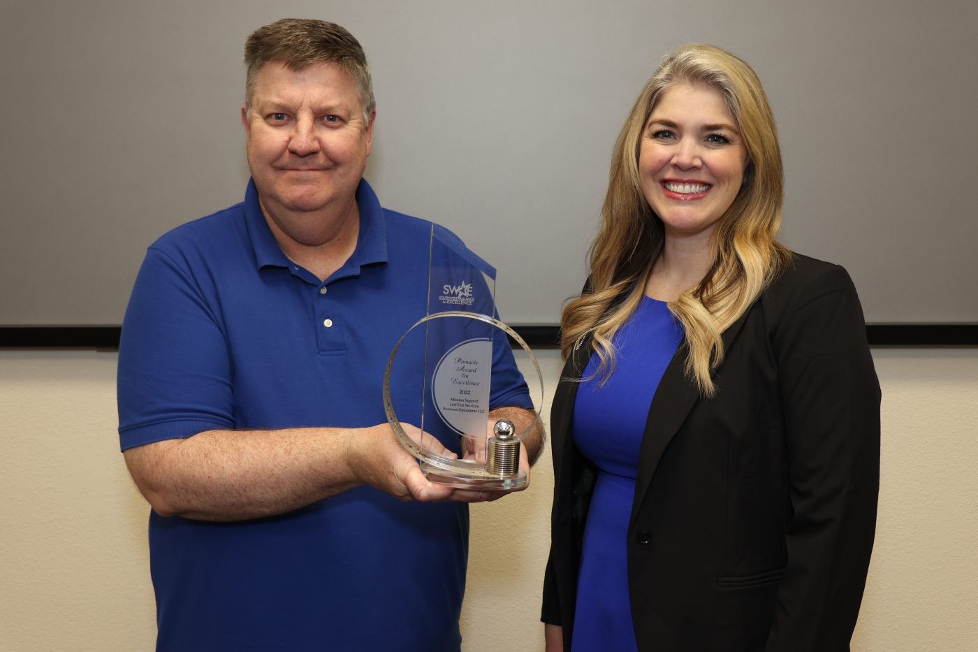 man on left in blue shirt holding glass award with woman in blue shirt and black jacket on right