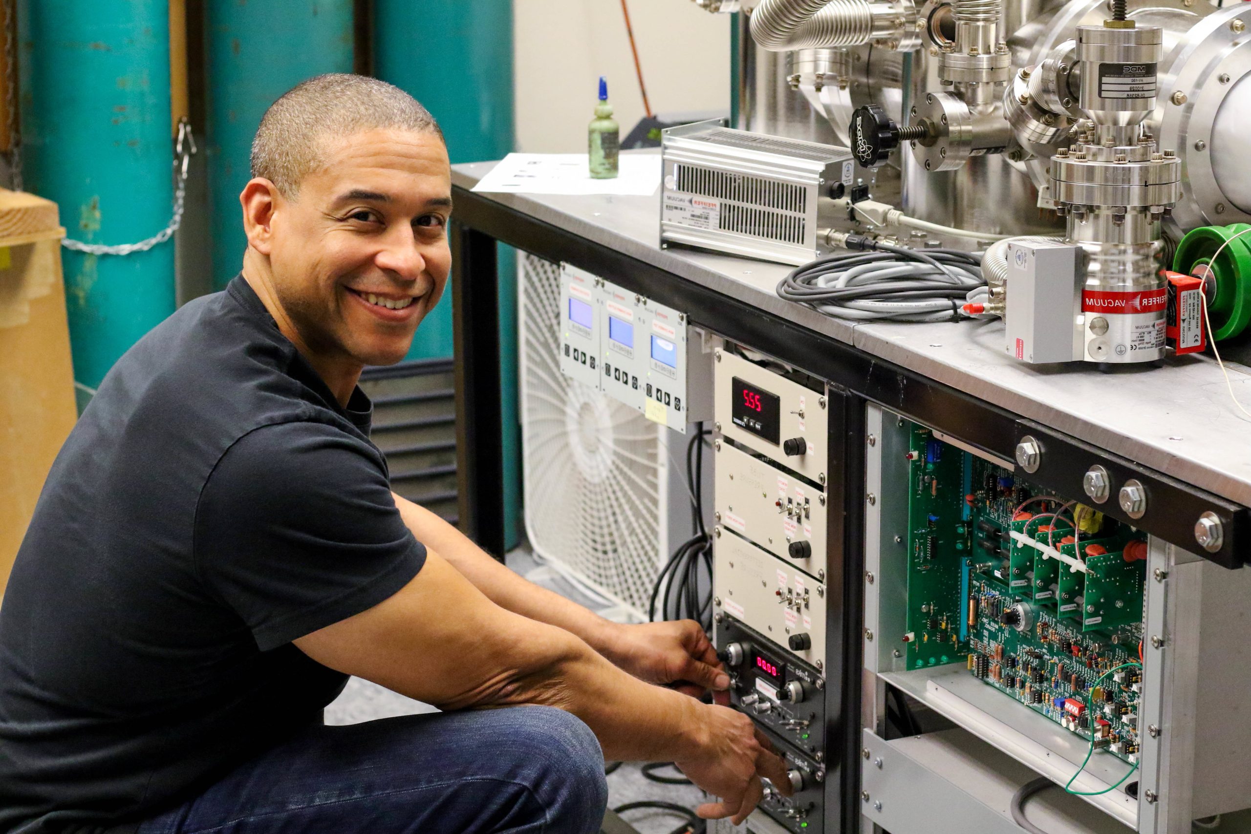 man on left in black t-shirt squatting in front of machinery and smiling at the camera