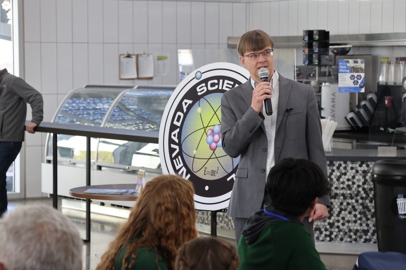 David Bowman in gray jacket standing and talking into microphone in a cafeteria