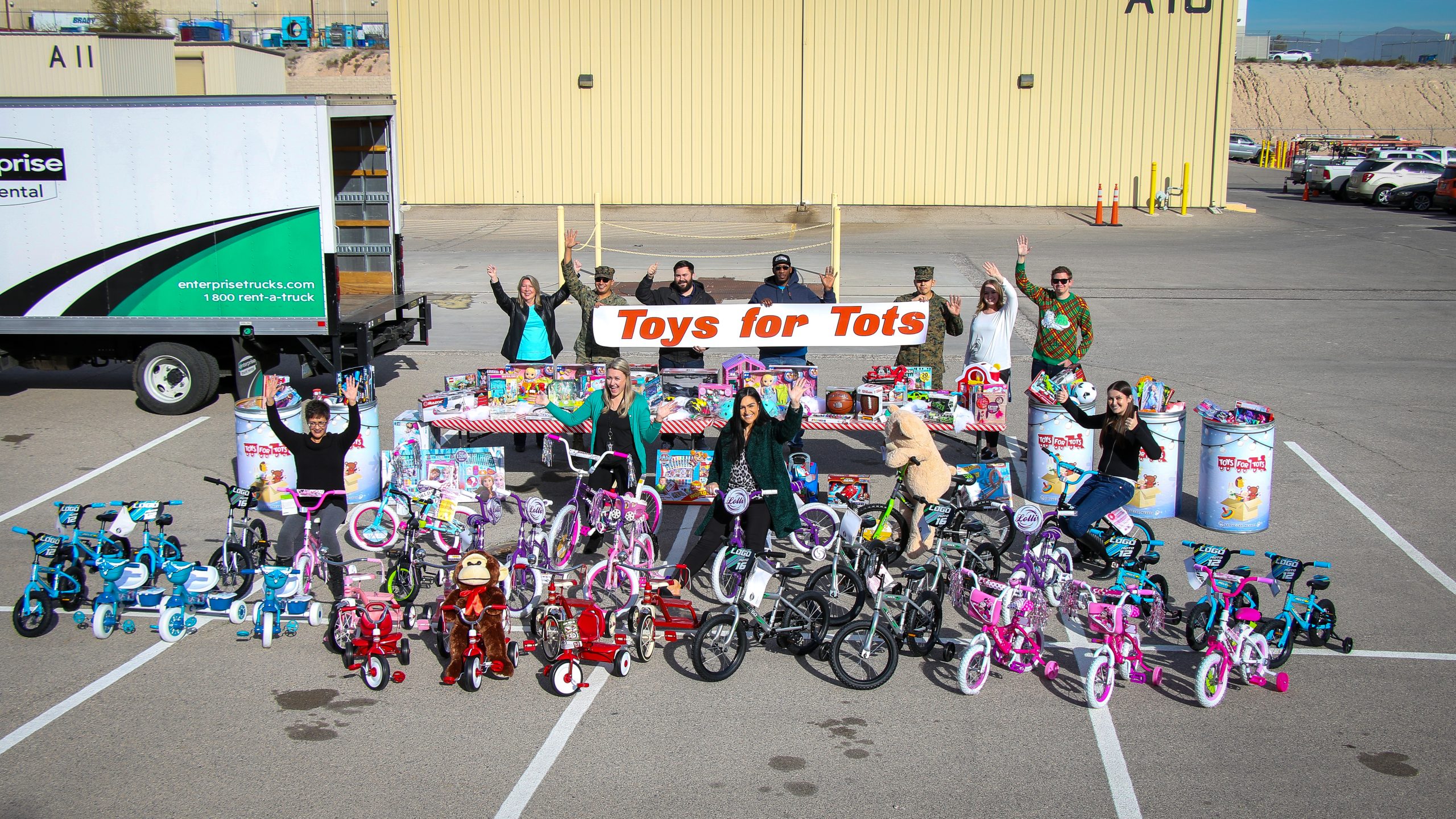 Bicycles and toys displayed in parking lot with people waving and a truck to the left