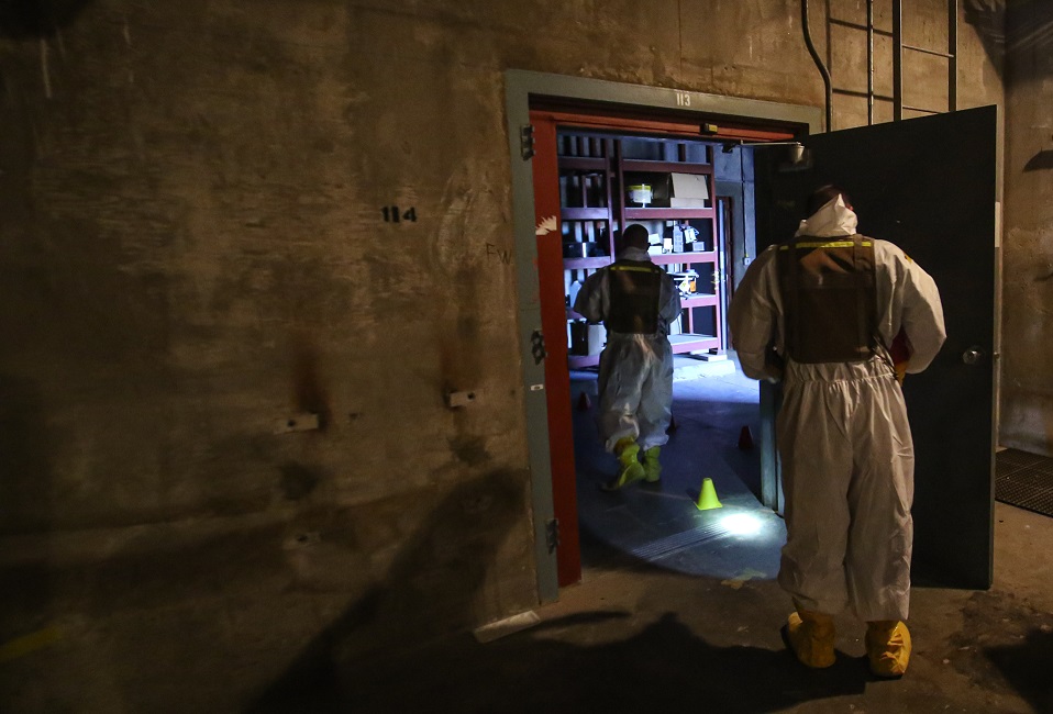 Two trainees in personal protective equipment enter a training facility using radiological detection equipment.