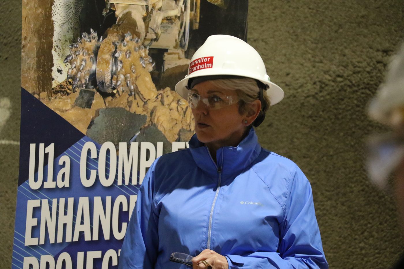 women in blue jacket and white hard hat with a red sticker on it in front of U1a Complex Enhancement Project banner
