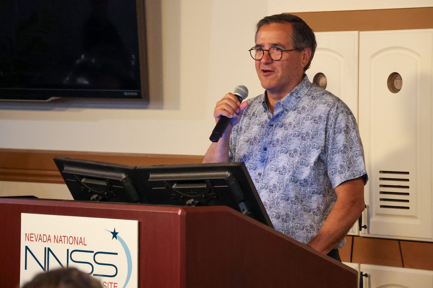 man in blue and white shirt standing at a podium speaking into a handheld microphone