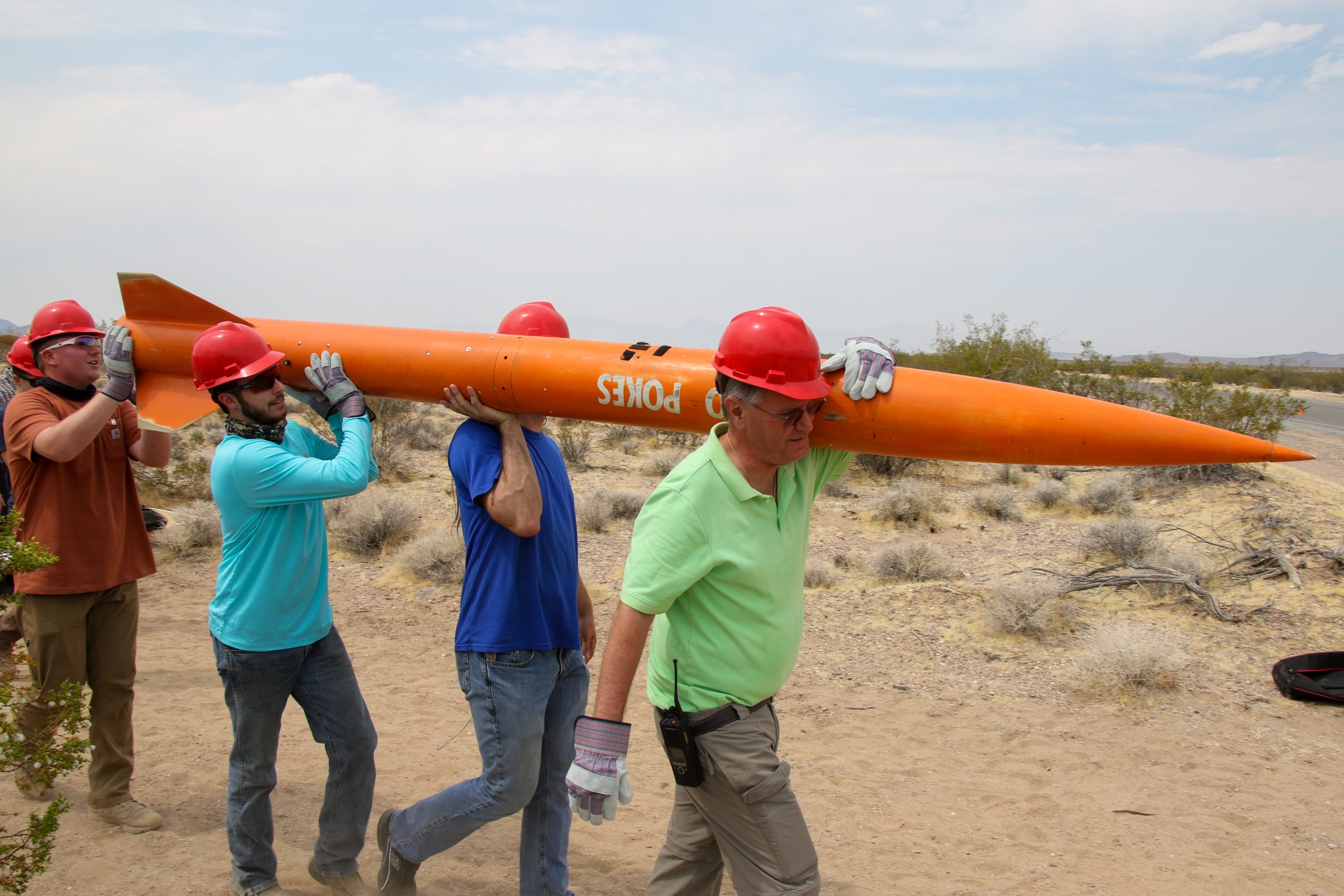 four men in red hard hats carrying an orange rocket in the desert