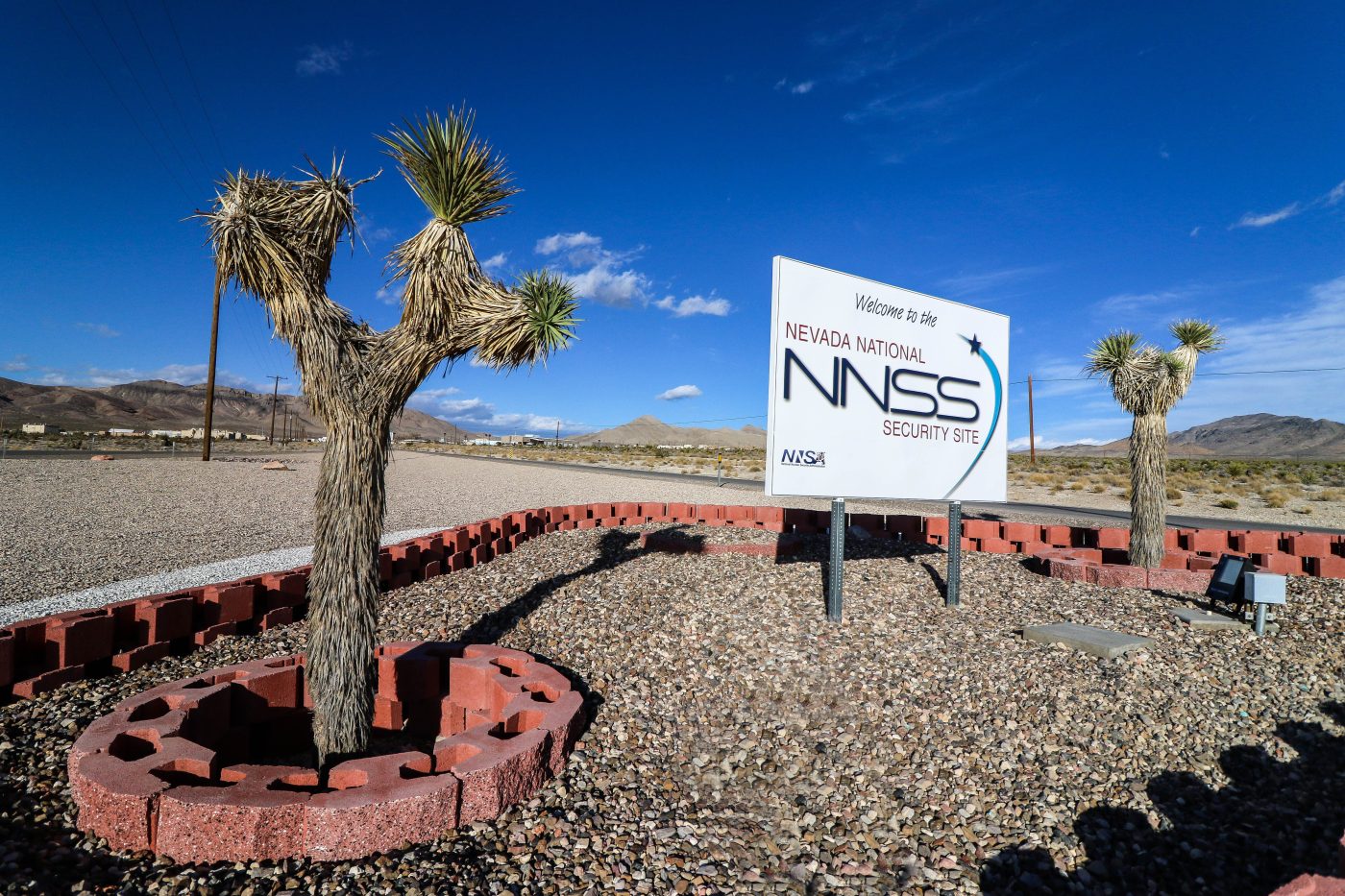 white sign that says welcome to the NNSS in a red bed with two joshua trees around it