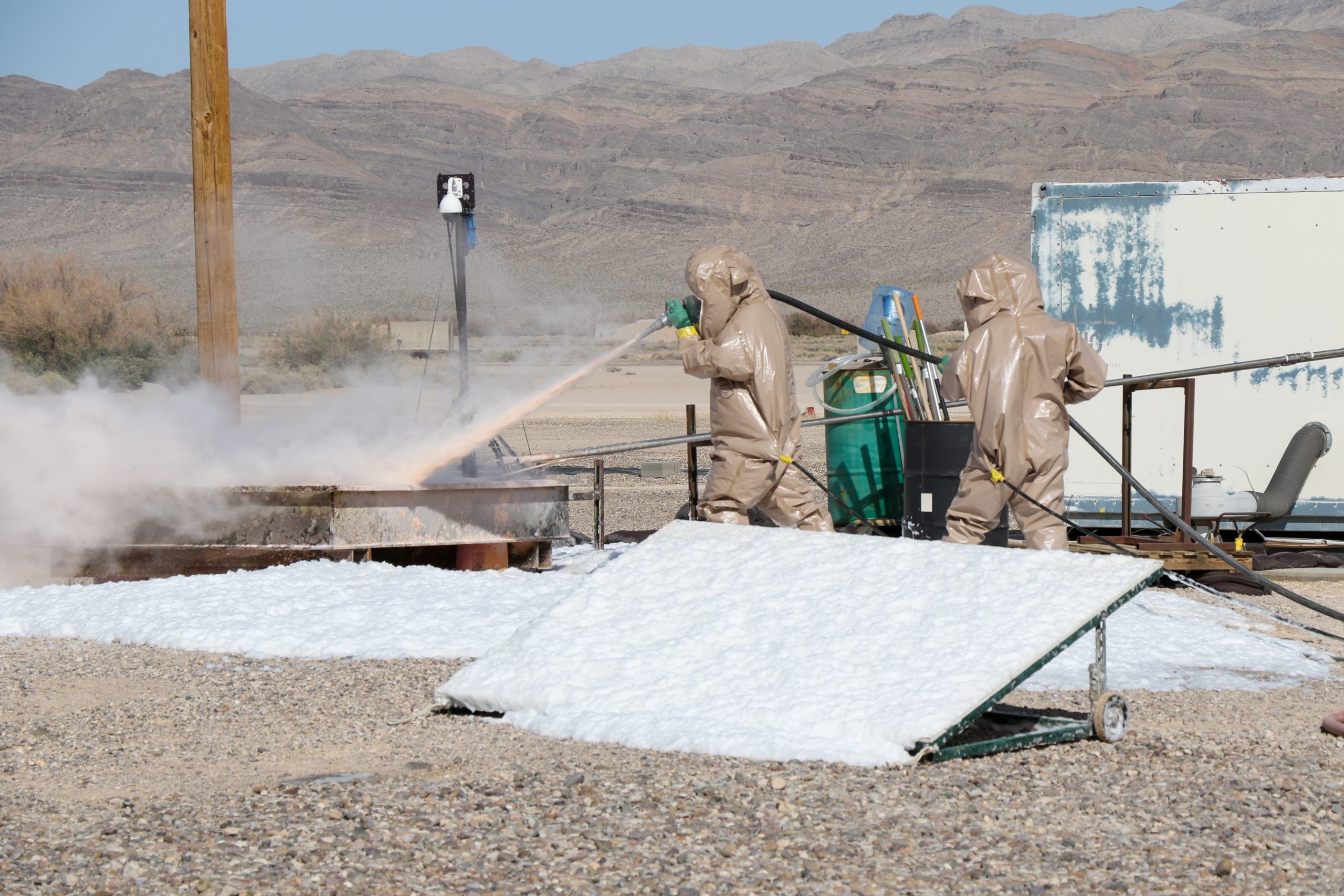 Two individuals in person protective equipment suits respond to a test bed experiment.