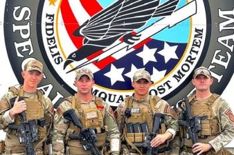 Four men in camouflage uniforms stand in front of a logo featuring an eagle and the United States flag.