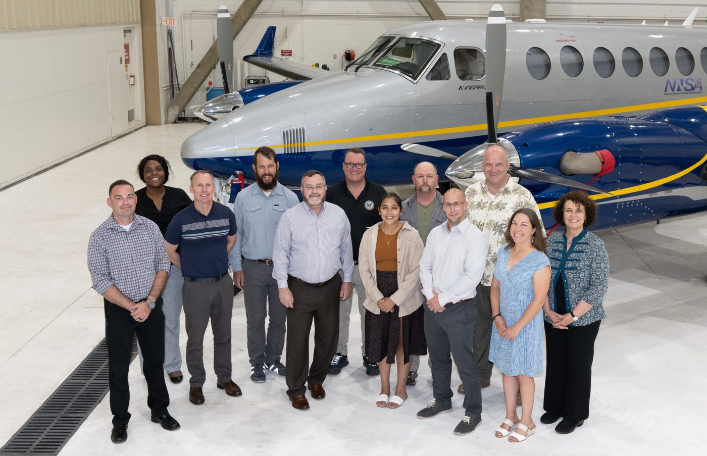 Twelve smiling people stand in front of a aircraft inside an aviation hangar.