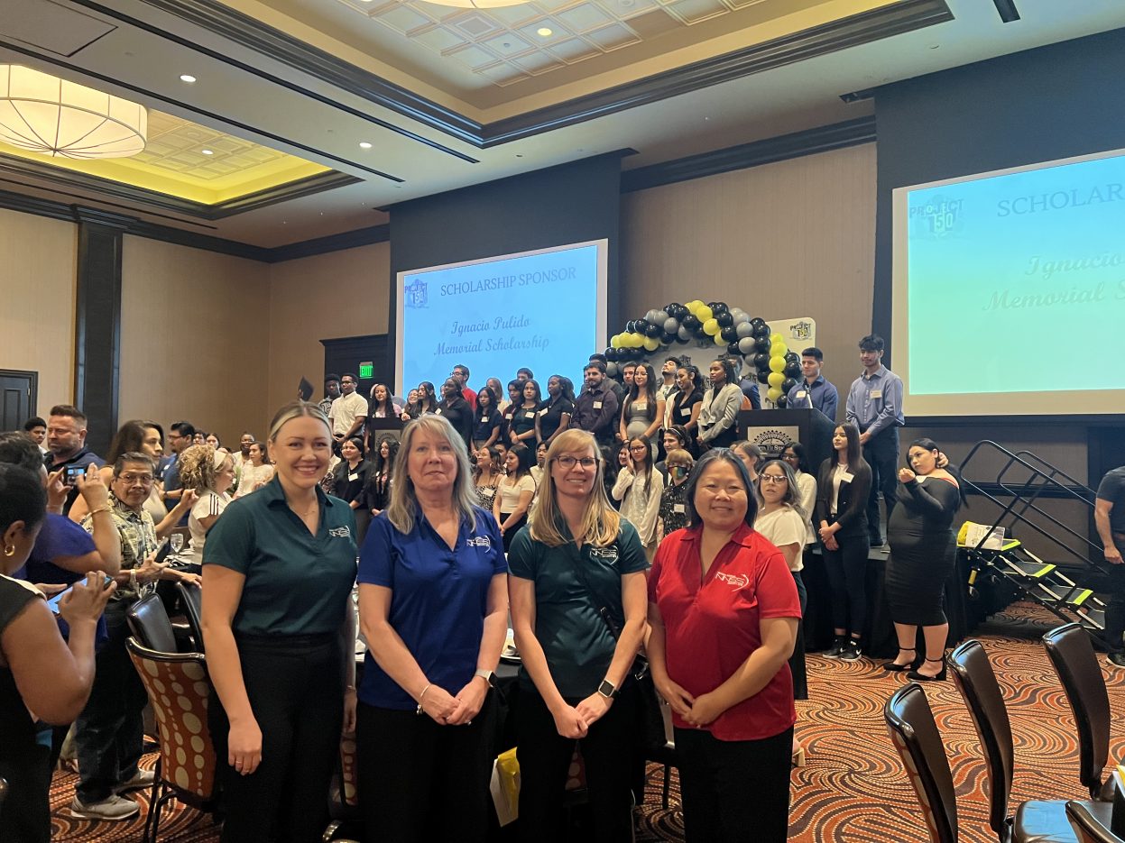 Four women in NNSS polo shirts pose for photo in ballroom with students on stage in the background