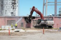 heavy machinery performing demolition on a pink building while two people in protective equipment spray water for dust mitigation