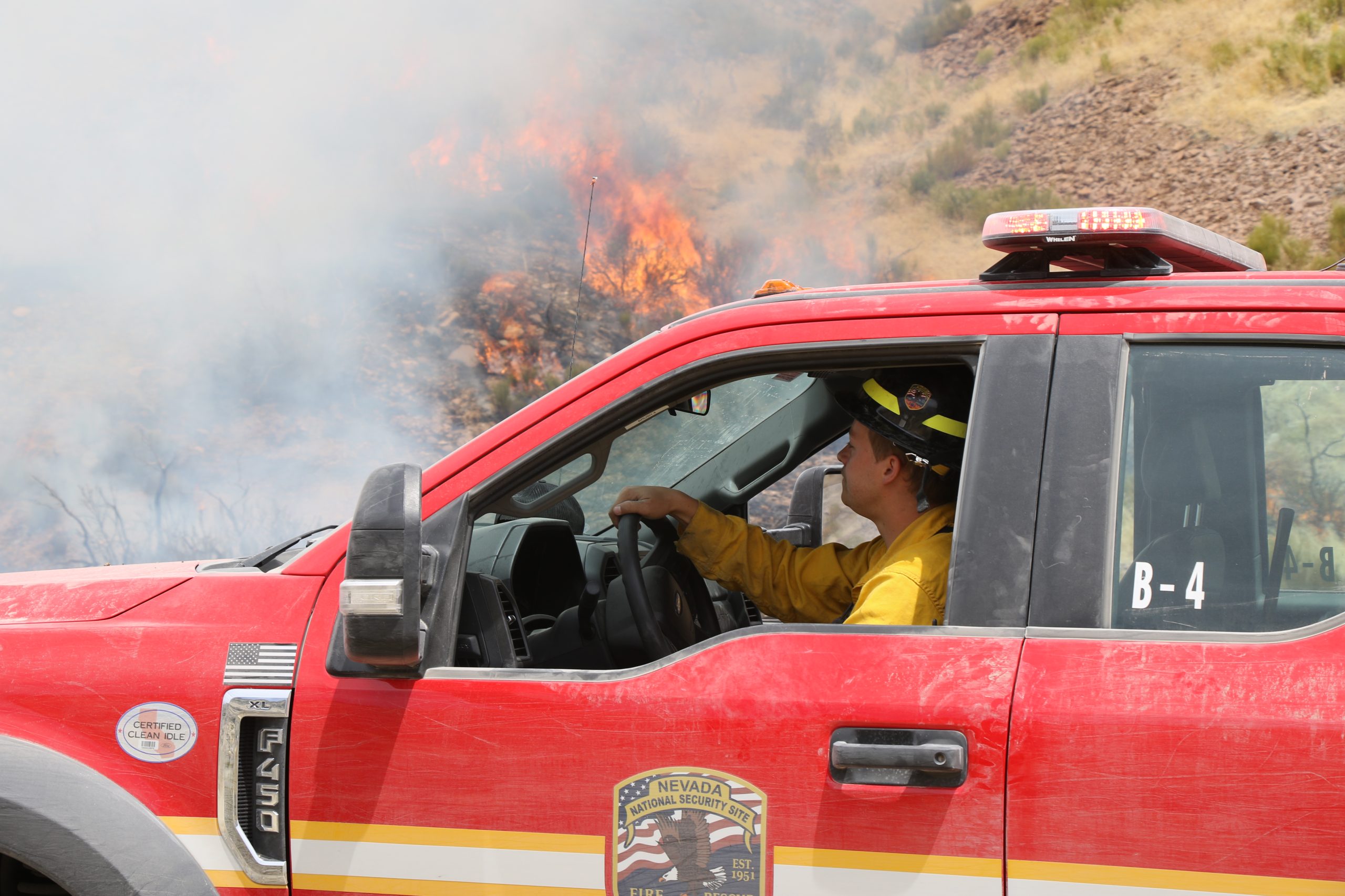 A firefighter drives a red vehicle near a wildland fire,