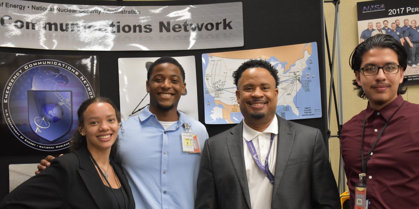 Three interns and a NNSS leader smile in front of Emergency Communications Network signage.