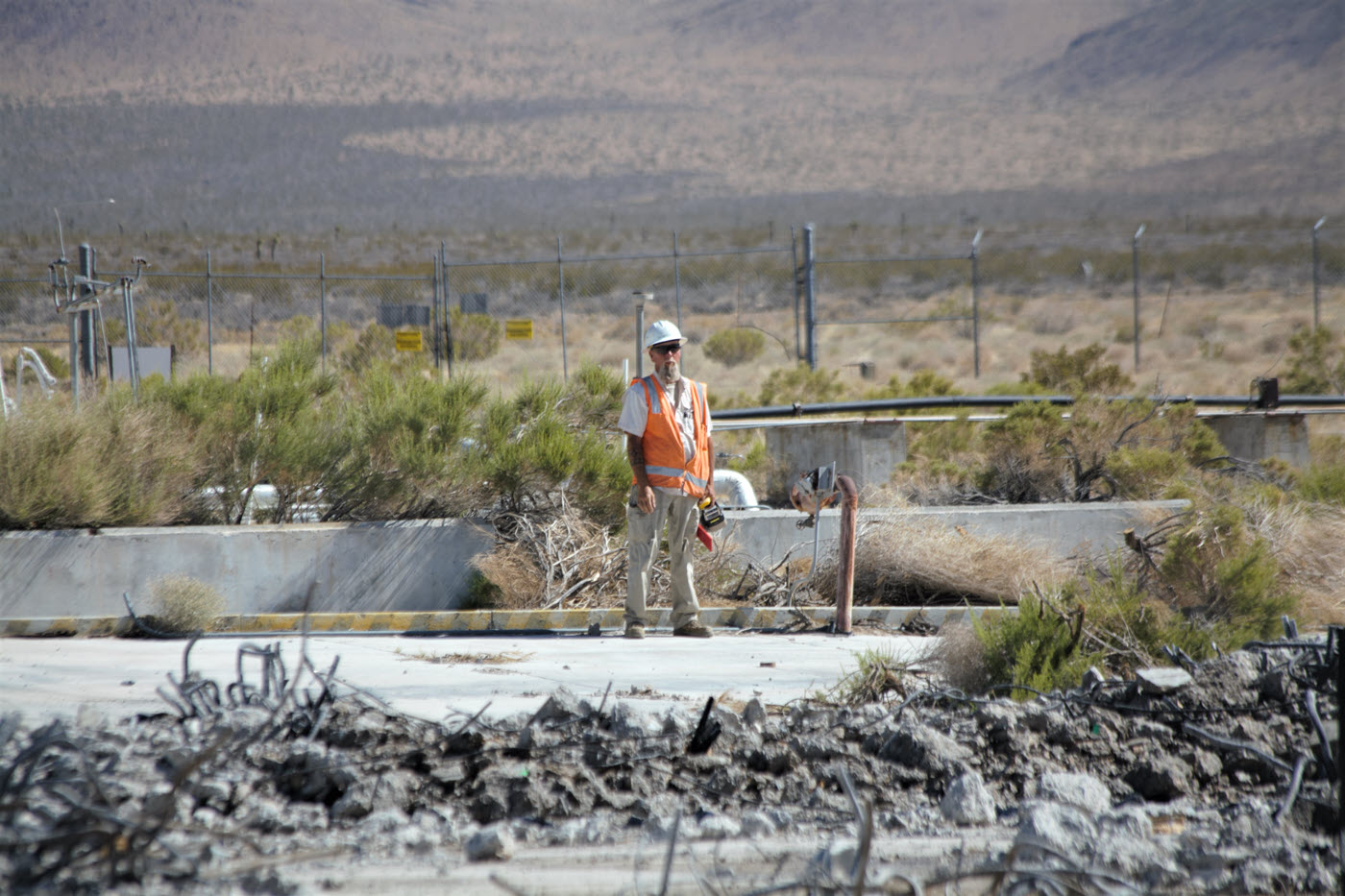 man in hard hat and orange vest standing on concrete pad surrounded by rubble in foreground and desert in background